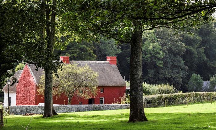 An old farmhouse has a thatched roof and two small chimneys. The exterior walls are painted red. there is a small tree in front of the farmhouse and t